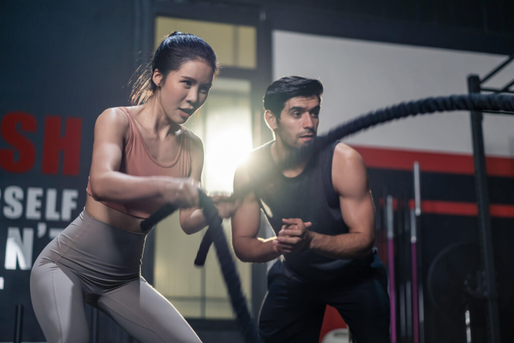 Trainer motivating a woman during battle rope training in a gym while discussing how long should your workouts be.